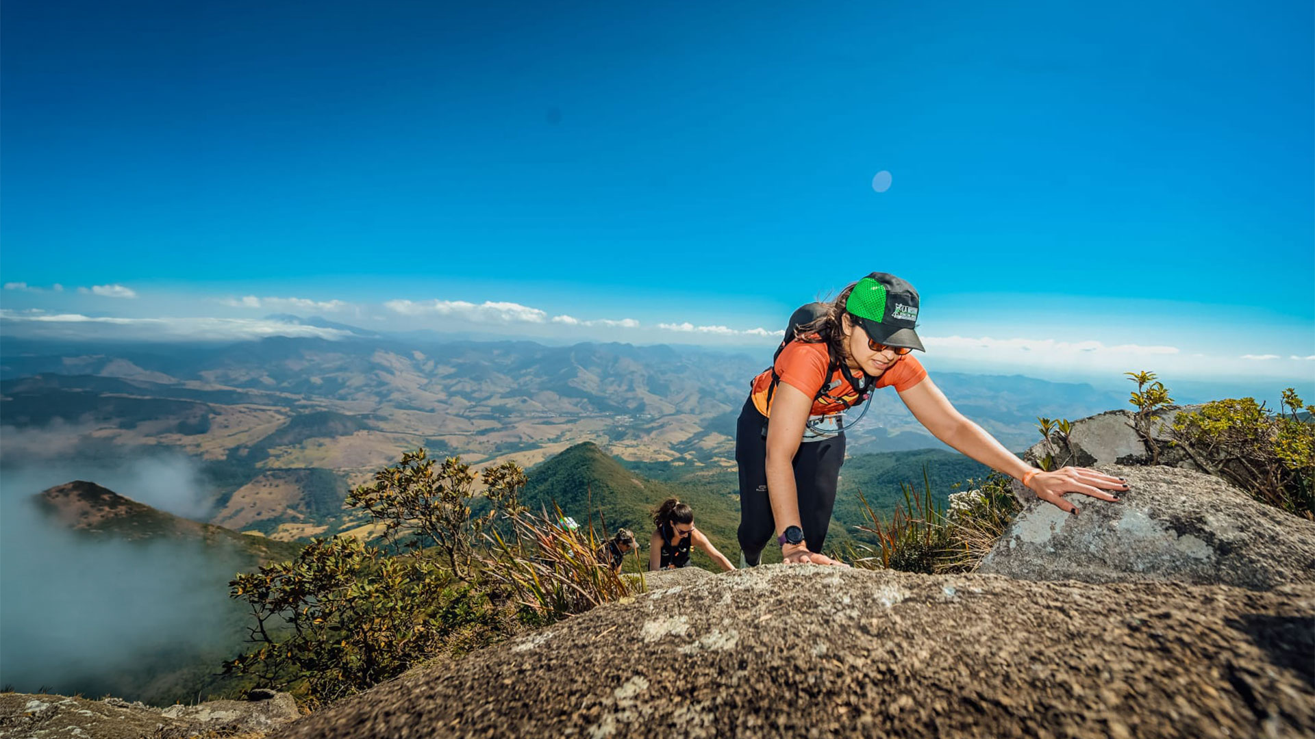 Corredora da Cerrado Trail escalando o costão de pedra de um cume, com a serra e o vale ao fundo