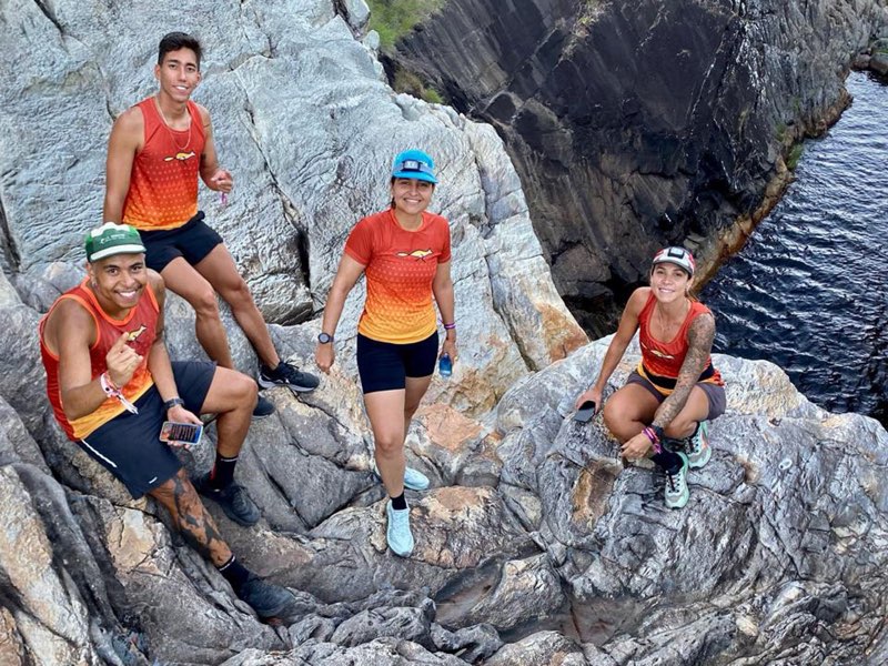 Quatro atletas de camisa laranja do clube sentados num costão de pedra à beira de um rio