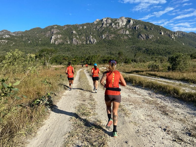Grupo do clube correndo numa estrada de terra em direção a uma serra rochosa sob céu azul