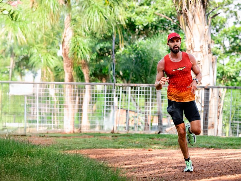 Atleta de regata laranja e boné vermelho correndo num caminho de terra dentro de um parque arborizado